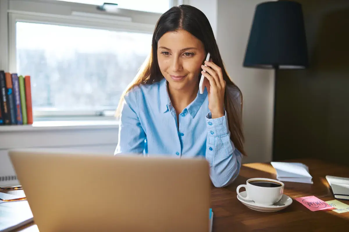 Woman on phone working at laptop with coffee