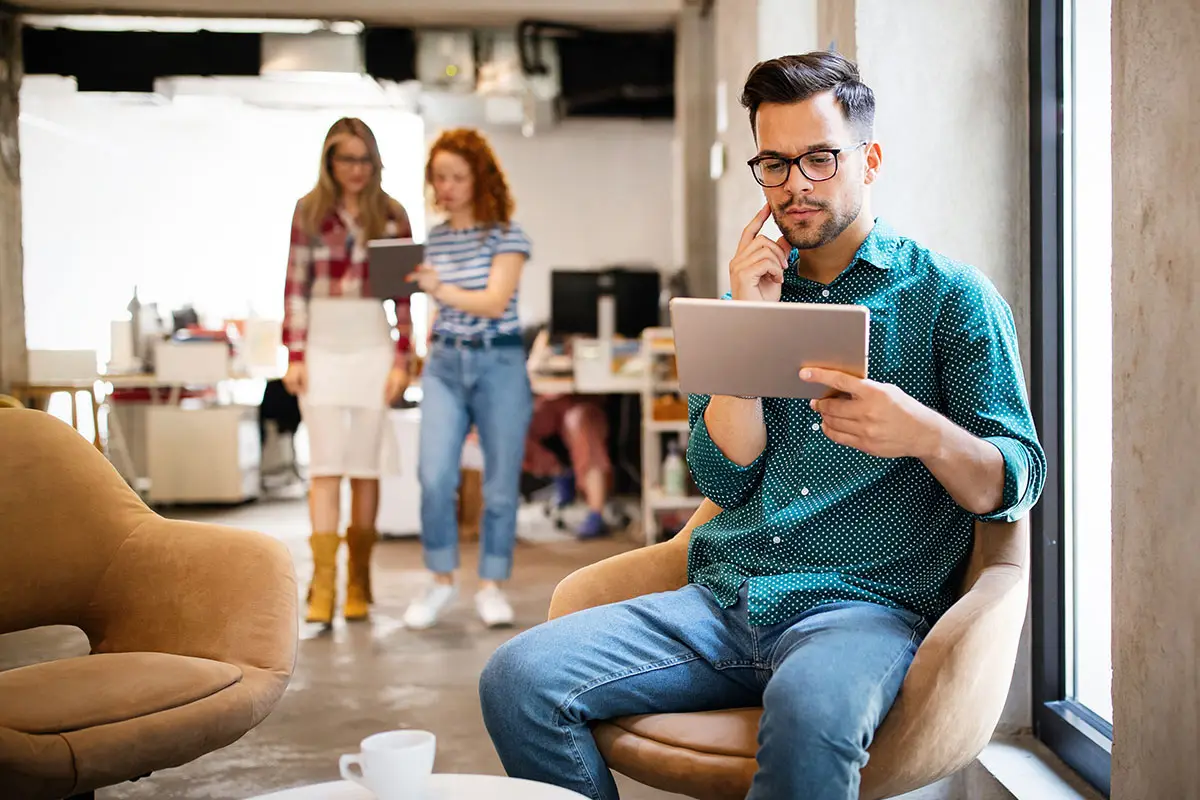 A man wearing glasses sits in a casual office checking his website domain expiration date on an ipad.